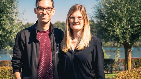 Appartementhotel Seeblick: Bildergalerie Junges Paar mit Brille steht draußen vor Bäumen und Wasser bei sonnigem Wetter