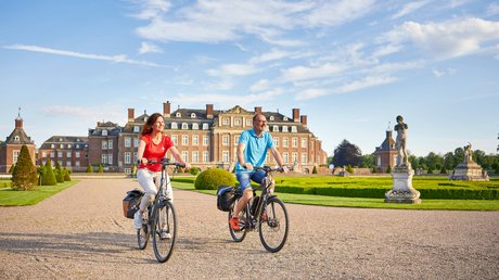Appartementhotel Seeblick: Bildergalerie Zwei Personen fahren mit dem Fahrrad vor einem Schloss und gepflegtem Garten