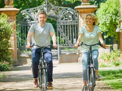 Seeblick: holidays by the lake in North Rhine-Westphalia Smiling couple riding bicycles on a sunny path with greenery and gate in background