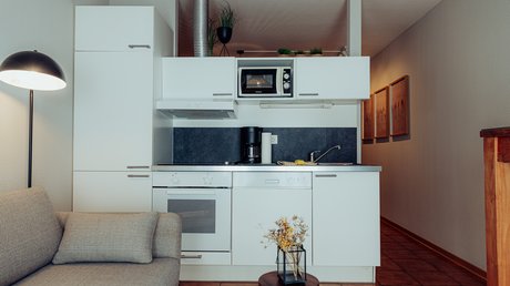 Appartementhotel Seeblick: Bildergalerie Kleine moderne Küche neben einem Sofa mit Stehlampe und Tisch mit Blumendekor.