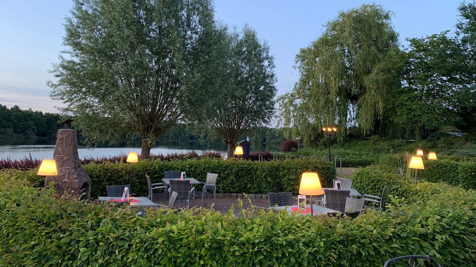 Hotel Seeblick in Gronau Abendliche Terrasse mit beleuchteten Tischen neben einem See und Bäumen