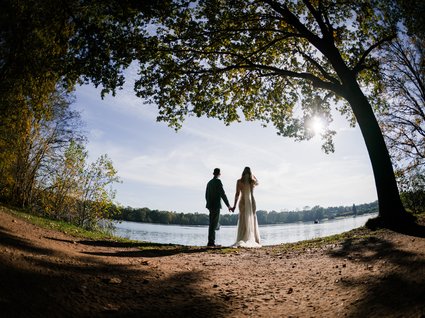 News vom Hotel direkt am See in NRW Paar in Hochzeitskleidung hält Händchen am See unter Bäumen bei Sonnenschein